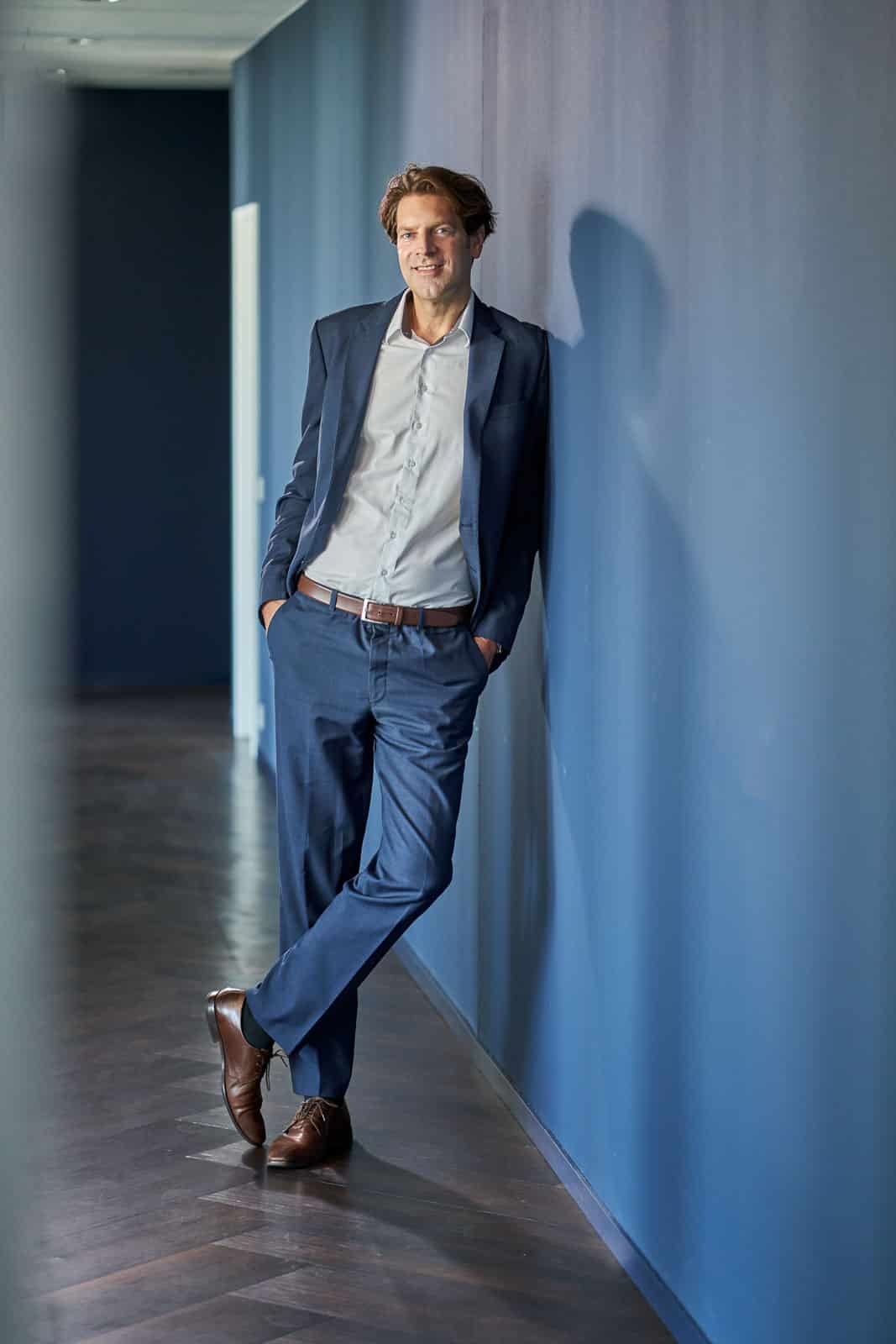 Home 23 Business-Portrait eines Mannes im Anzug, der an einer blauen Wand lehnt.